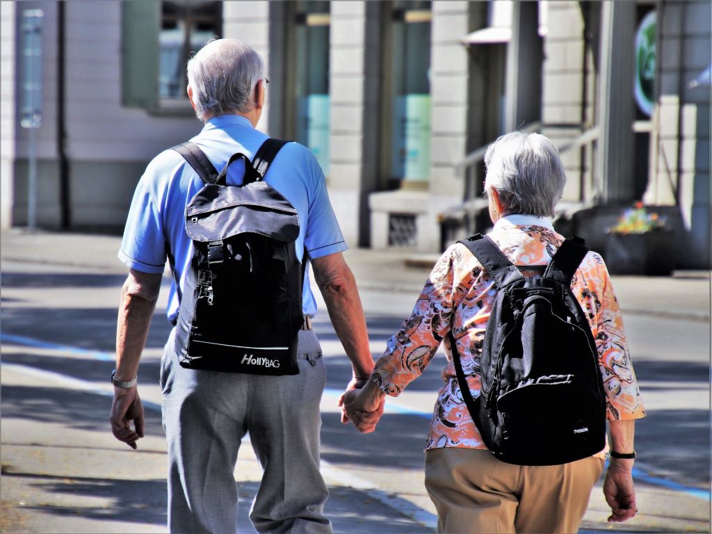 An older man and woman hold hands and carry bags.
